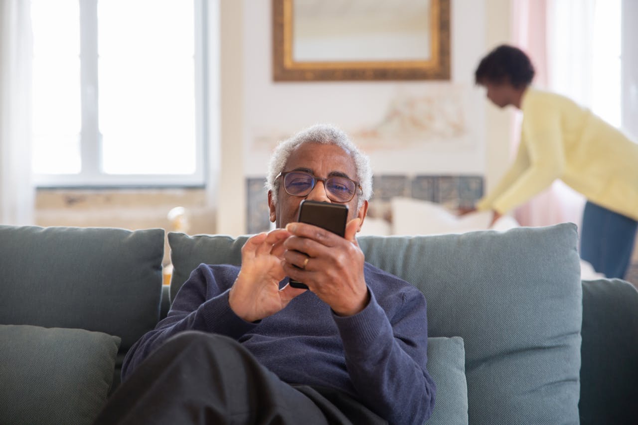 Elderly man sitting on couch using smartphone indoors. Cozy home setting.